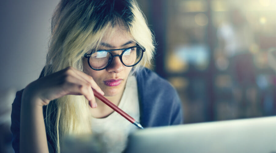 a woman with glasses holding a pen whilst browsing on her laptop