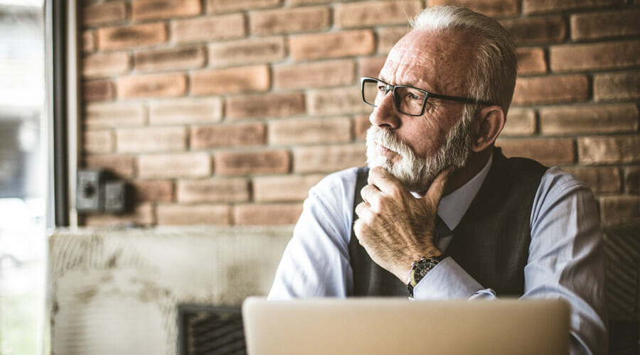 a smartly dressed elderly gentleman with glasses pondering whilst on his laptop