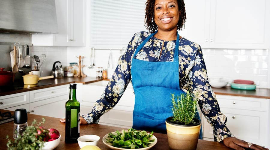 a woman wearing an apron in her kitchen, smiling at the camera. In front of her on the counter are vegetables, herbs and olive oil.