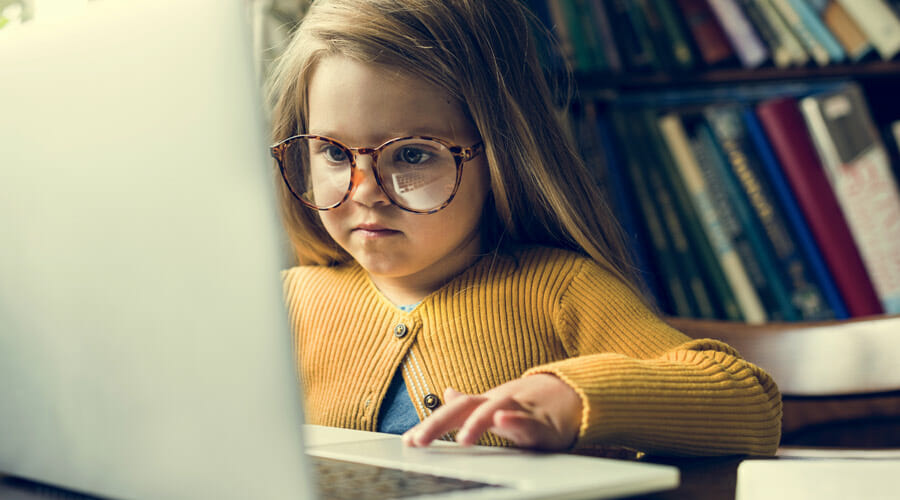 a child with glasses sat at a desk whilst using a laptop. a bookshelf is in the background.