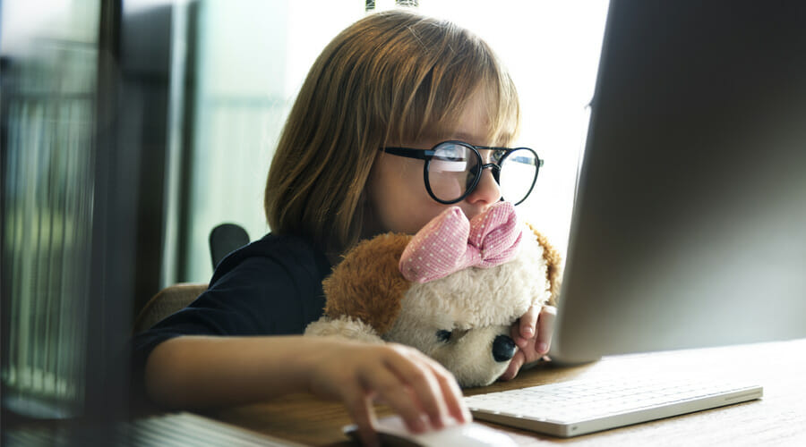 A child with glasses sat at a desk whilst leaning on her stuffed animal. She staring intently at an Apple computer.