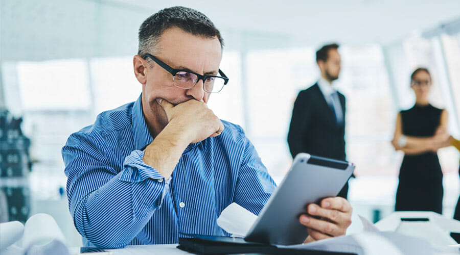 a middle-aged man with glasses is sat at a desk, using a tablet. He is staring intently at the tablet whilst his hand overs his mouth