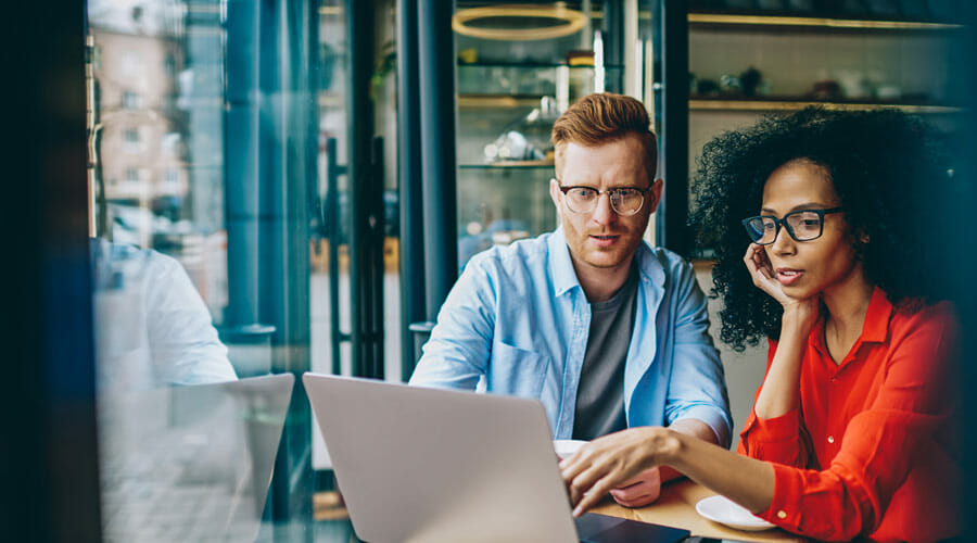 a man and a woman with glasses are sat in a cafe whilst looking at a laptop