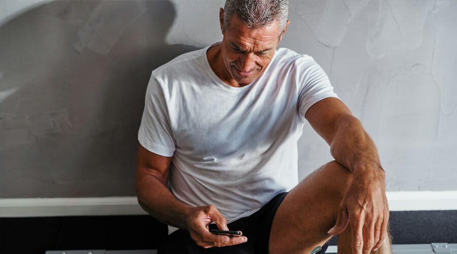 An older man sat against a wall whilst looking down and scrolling on his phone