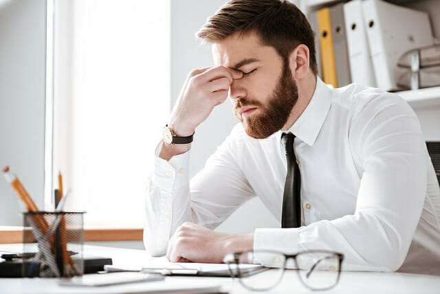 a man holding the bridge of his nose in pain whilst sat at his desk