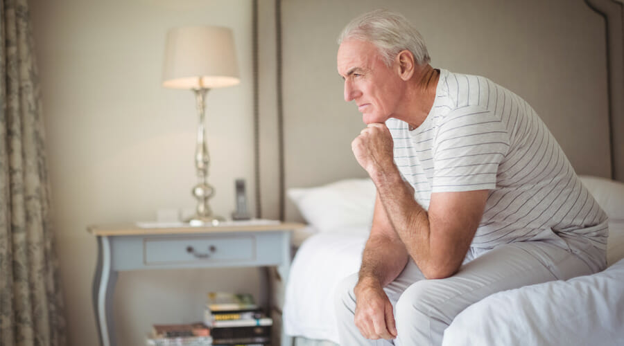 an older gentleman sitting on the edge of his bed in deep thought