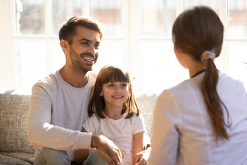Step-2-children-samer-hamada-eye-clinic-london a little girl and her father at an eye clinic appointment. They sat on a sofa smiling whilst talking to a medical professional.