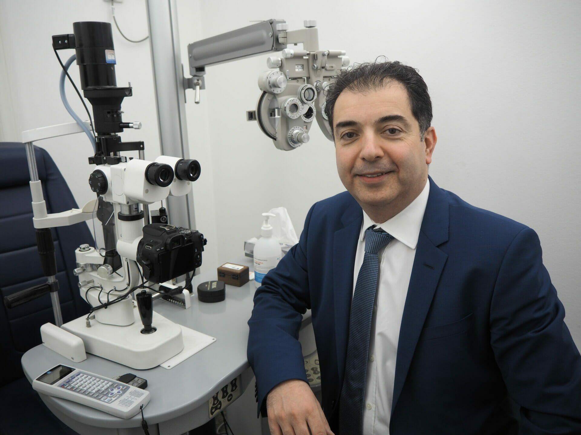 A portrait photo of Mr Samer Hamada smiling in a suit whilst leaning on a desk at the forefront of the image. In the background you can see eye testing equipment.